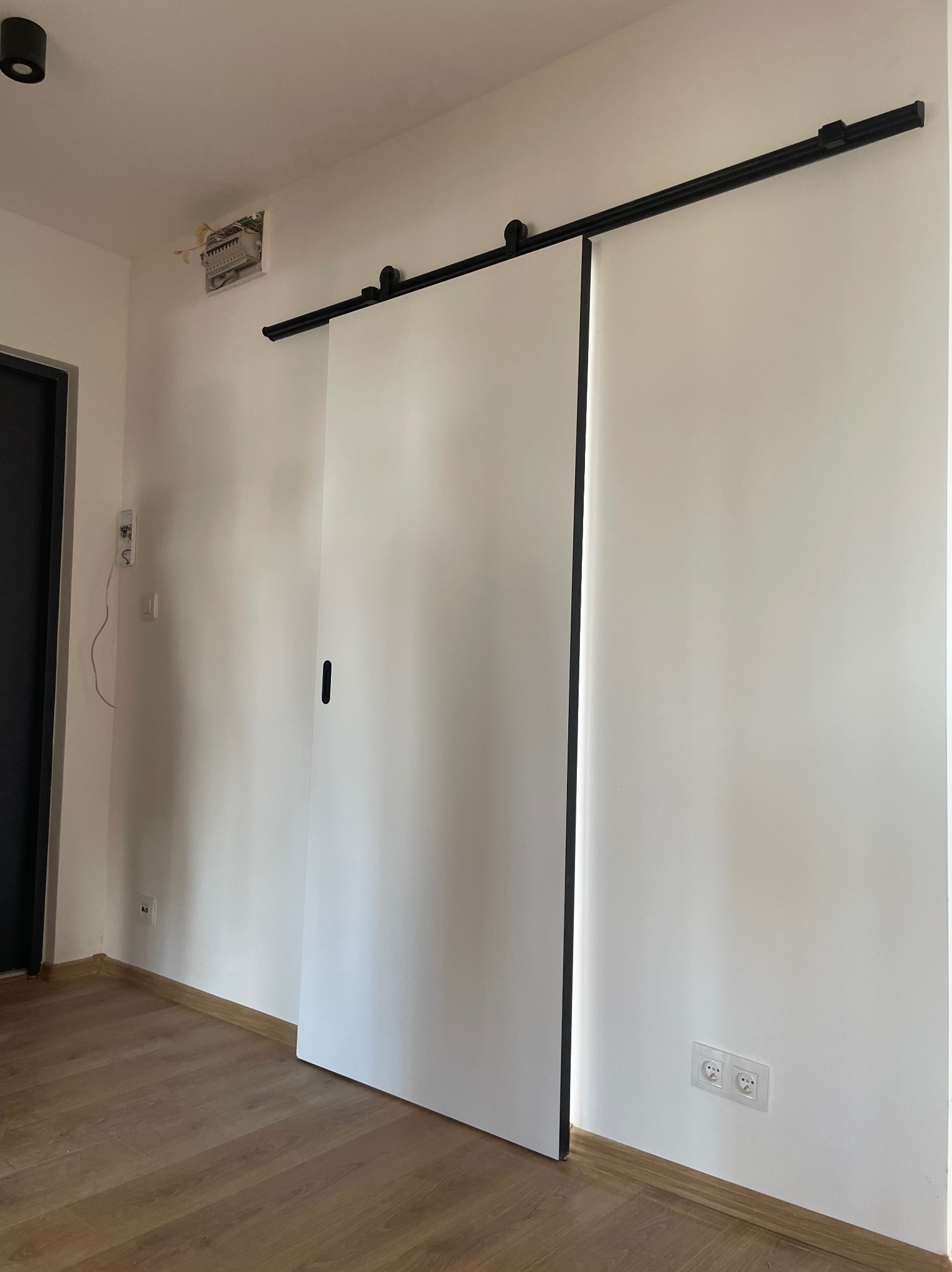 Białe przesuwne drzwi zamontowane na czarnej szynie, widoczne na tle białej ściany i drewnianej podłogi, z widocznymi gniazdkami elektrycznymi i otwartą puszką elektryczną w górnym rogu.