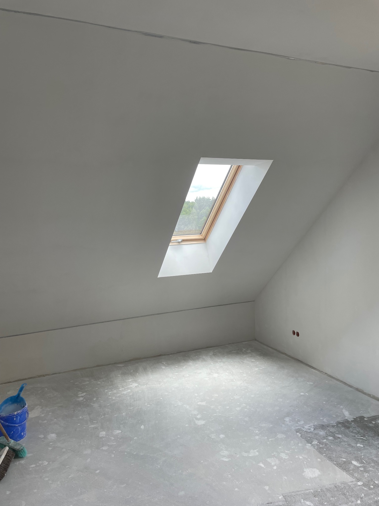 Poddasze po nałożeniu gładzi, widoczne ślady po pracy na podłodze, niebieskie wiadro z narzędziami w rogu, okno dachowe wpuszczające światło.