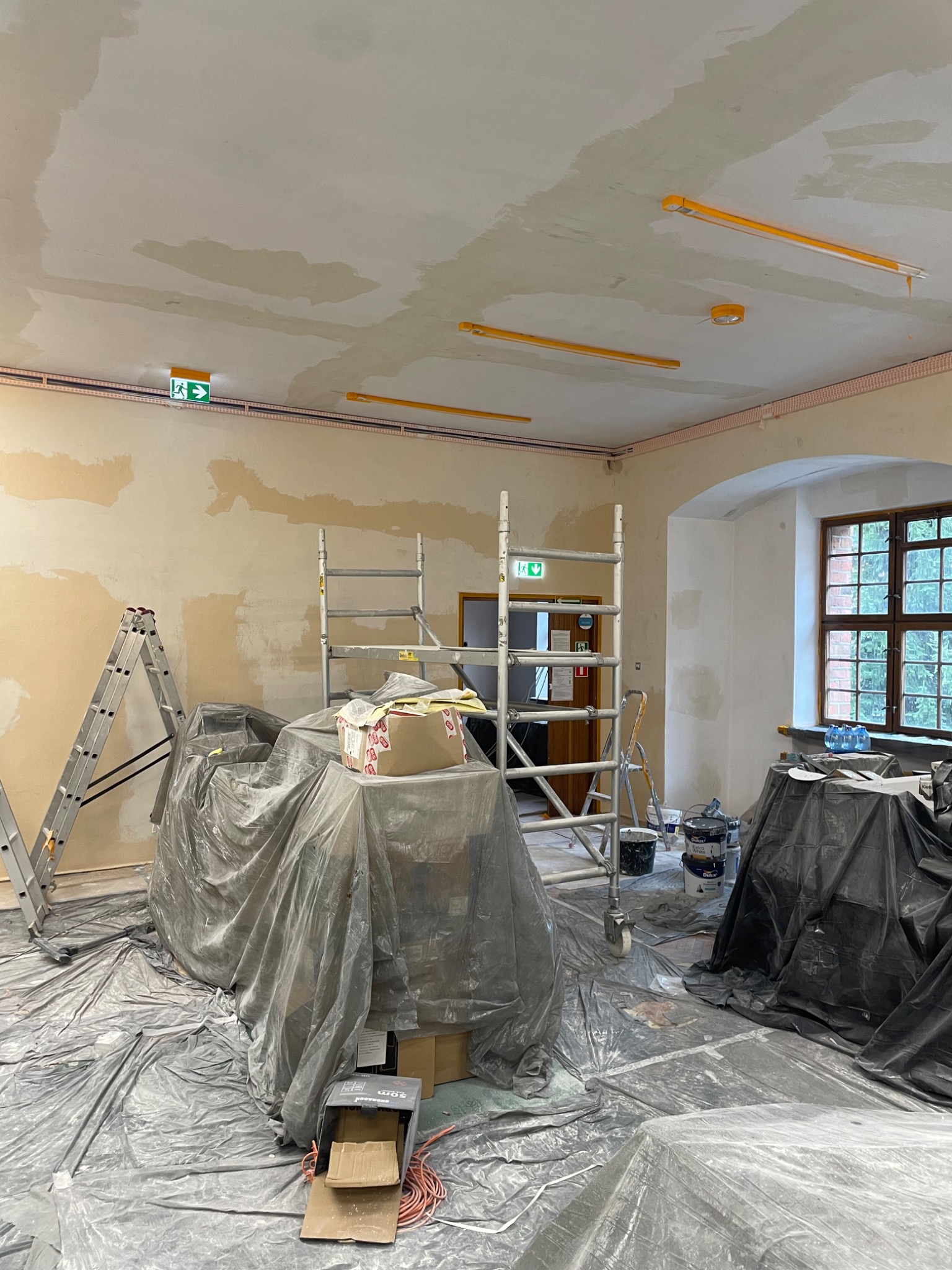 Wnętrze w trakcie prac przygotowawczych: ściany częściowo pokryte gładzią, rozstawione drabiny i rusztowania, podłoga zabezpieczona folią malarską, widoczne puszki z farbą i narzędzia.