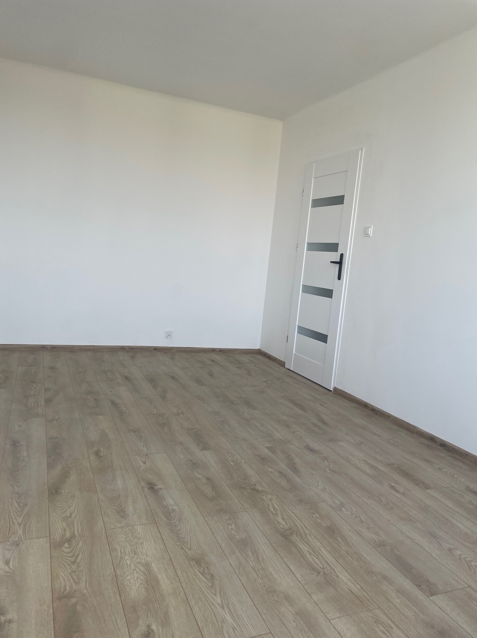 Pusty pokój po remoncie: świeżo ułożone panele podłogowe w odcieniach beżu i szarości, białe ściany i białe drzwi z czarną klamką.