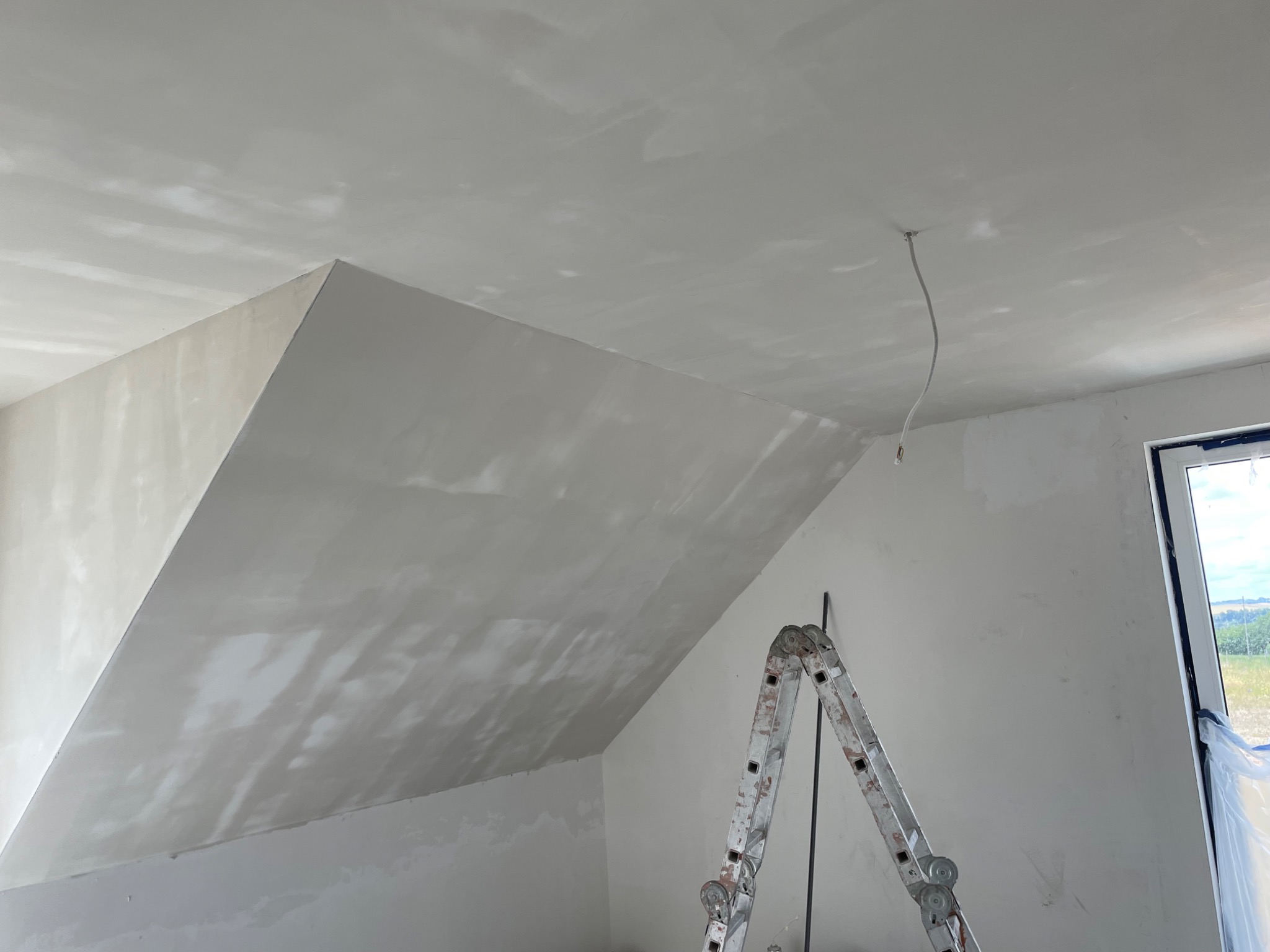 Poddasze po nałożeniu gładzi szpachlowej, widoczny fragment okna, kabel zwisający z sufitu oraz aluminiowa drabina oparta o ścianę.