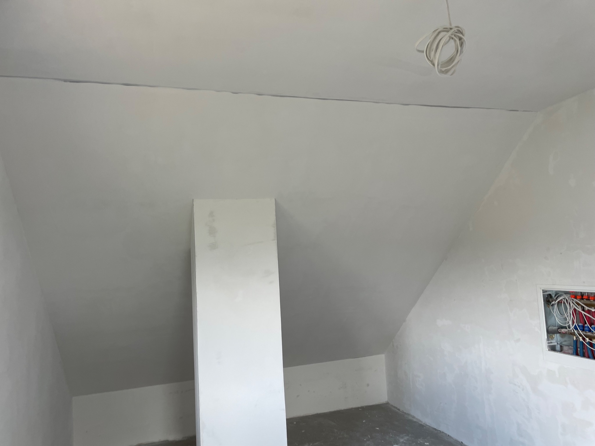 Poddasze w trakcie prac wykończeniowych, widoczna surowa ściana ze śladami szpachlowania, instalacja elektryczna zwisająca z sufitu i instalacja wodna w ścianie.