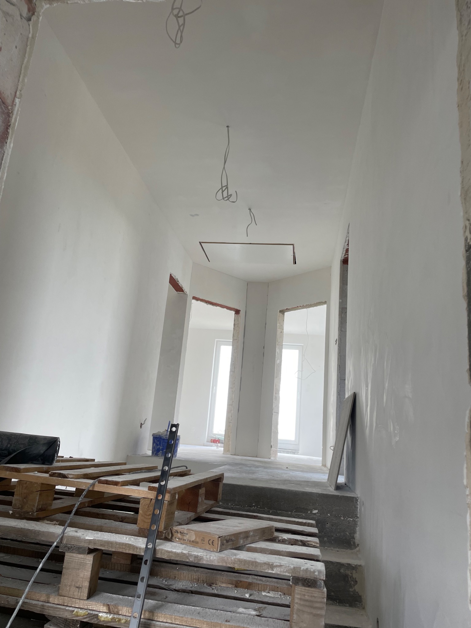 Wnętrze w trakcie prac wykończeniowych: świeżo pomalowane ściany na biało, widoczne kable zwisające z sufitu, palety na podłodze i surowy stan otworów drzwiowych.