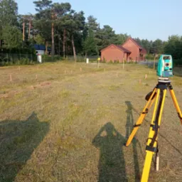 Tyczenie działki budowlanej z użyciem niwelatora optycznego na trójnogu, widoczne paliki geodezyjne w trawie i budynek mieszkalny w tle.