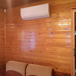 FM klima Sp. z o.o. - Biały klimatyzator marki Kaisai zamontowany na ścianie z boazerii w drewnianym pokoju, widoczny fragment sofy i przeszklona witryna z zastawą.