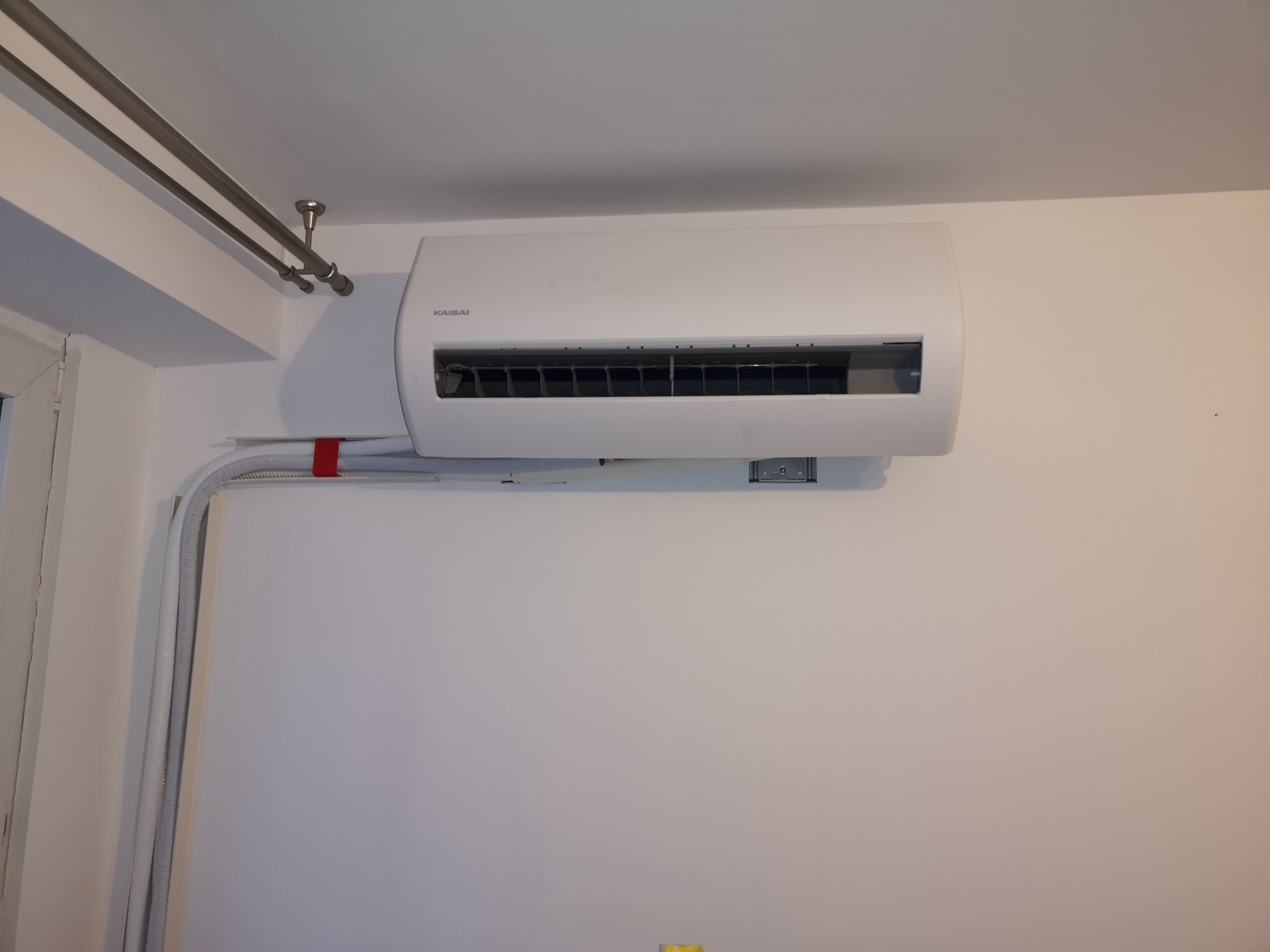 Biały klimatyzator Kaisai zamontowany na ścianie, widoczne rury instalacyjne i karnisz sufitowy