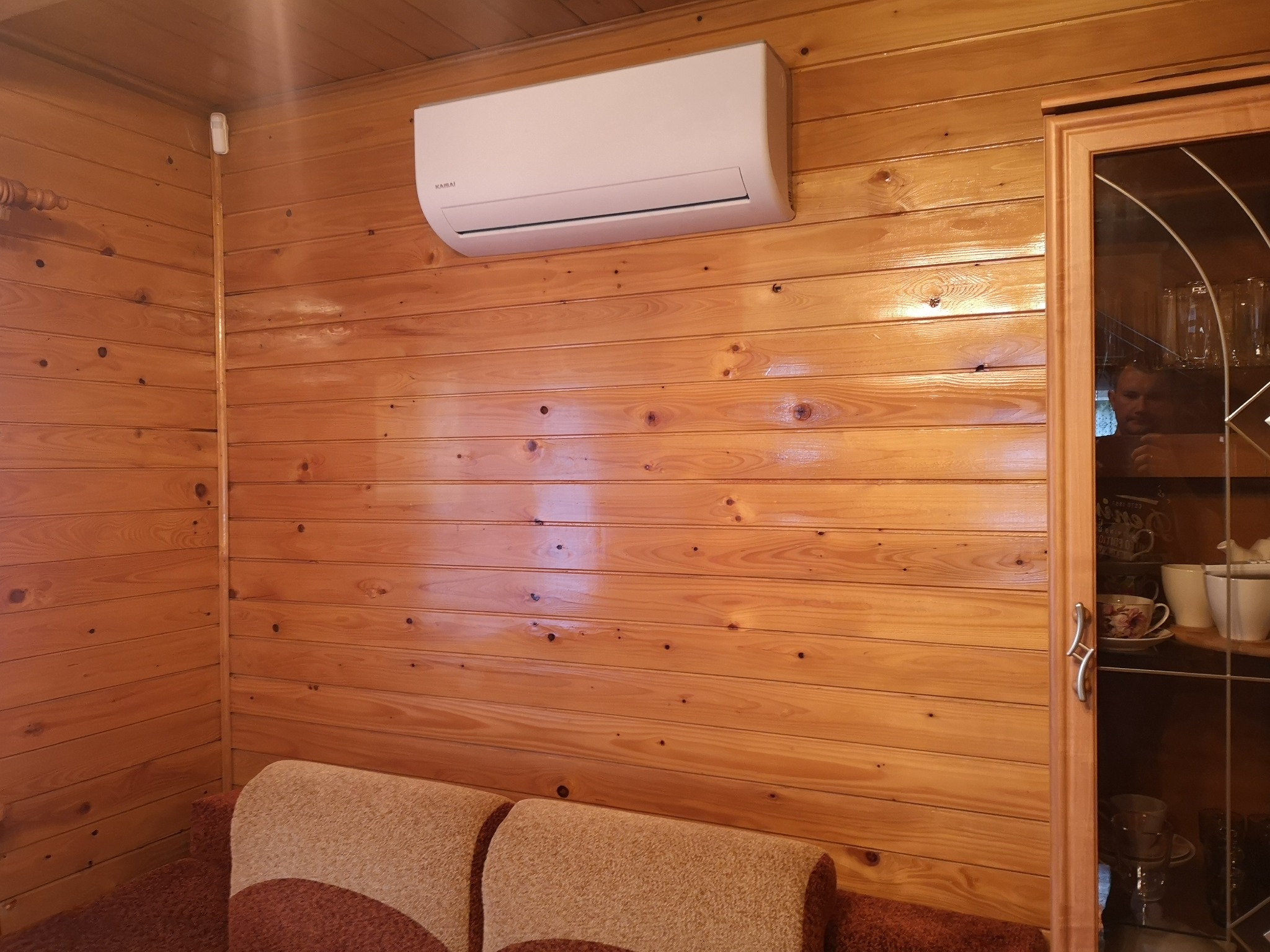 Biały klimatyzator marki Kaisai zamontowany na ścianie z boazerii w drewnianym pokoju, widoczny fragment sofy i przeszklona witryna z zastawą.