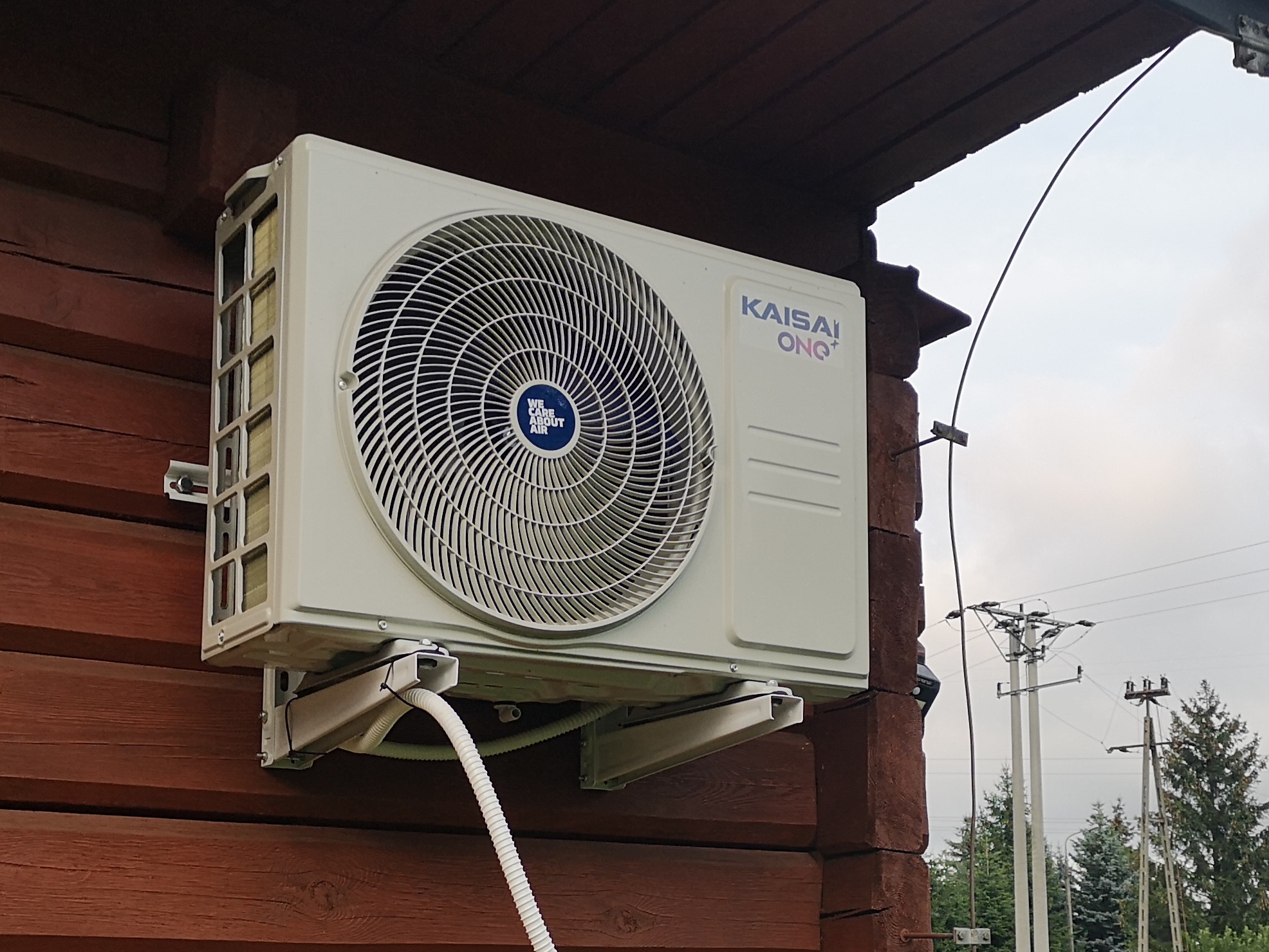 Biała jednostka zewnętrzna klimatyzatora marki Kaisai ONQ+ zamontowana na brązowej, drewnianej ścianie domu z widocznym przewodem i elementami montażowymi.