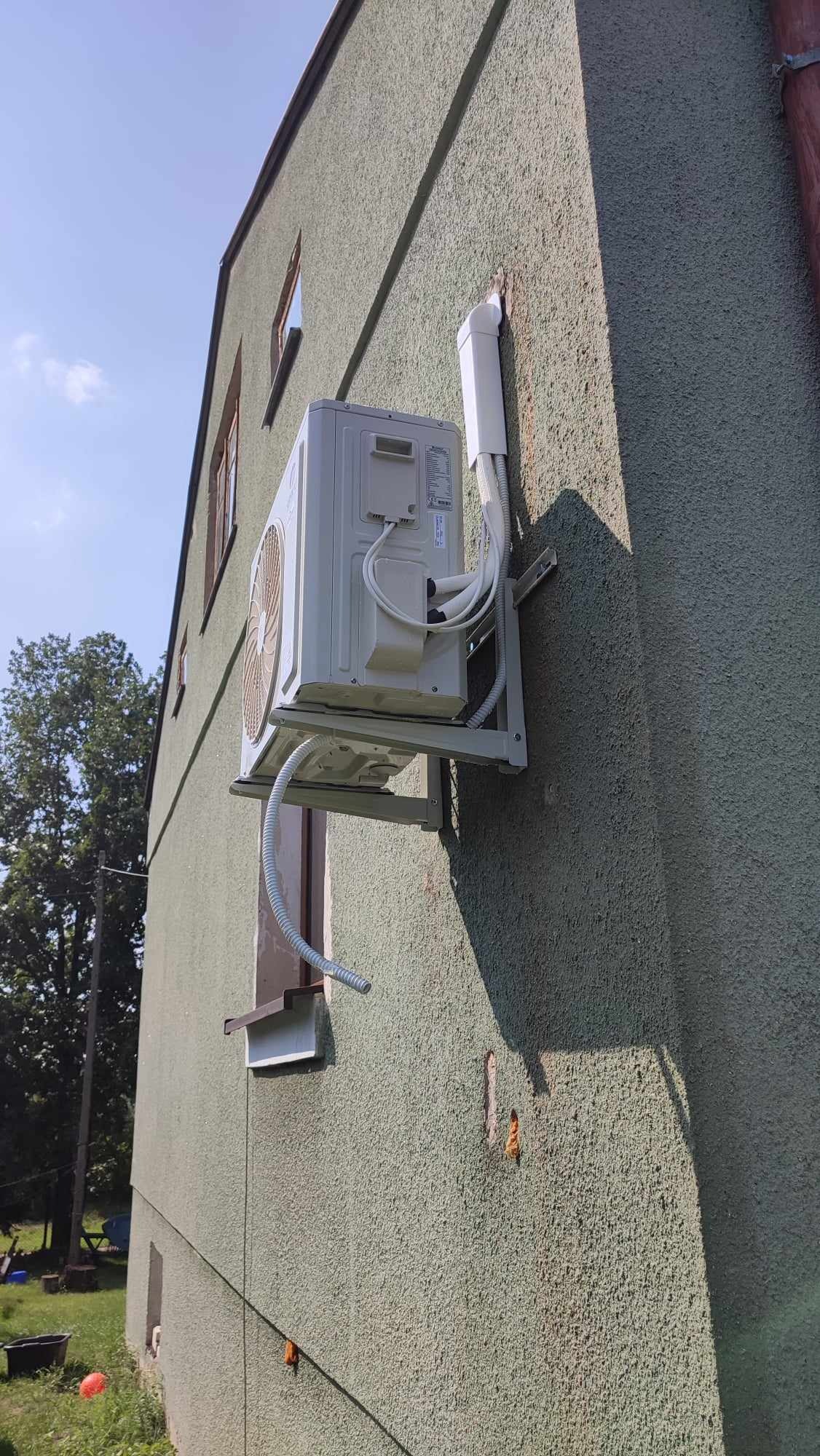 Zewnętrzna jednostka klimatyzacji zamontowana na ścianie budynku z zielonym, chropowatym tynkiem, widoczna instalacja odprowadzająca skropliny nad oknem.