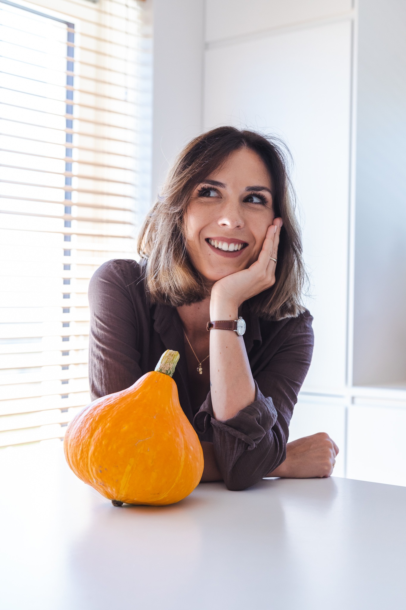 Uśmiechnięta kobieta z brązowymi włosami, opierająca brodę na dłoni, z zegarkiem i złotym łańcuszkiem, siedząca przy białym stole obok pomarańczowej dyni hokkaido.