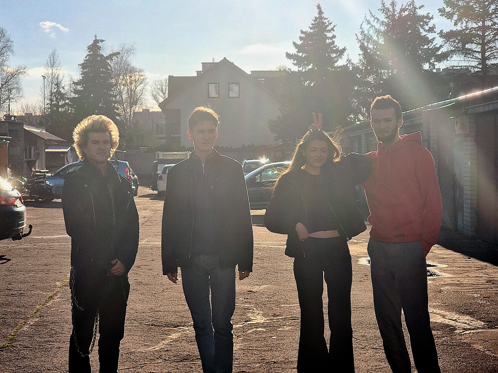 Czteroosobowa grupa młodych ludzi stoi na zewnątrz, na tle garaży i domu jednorodzinnego w słoneczny dzień. Dziewczyna z podniesionymi palcami tworzy uszy królika. Jeden z mężczyzn ma na sobie...