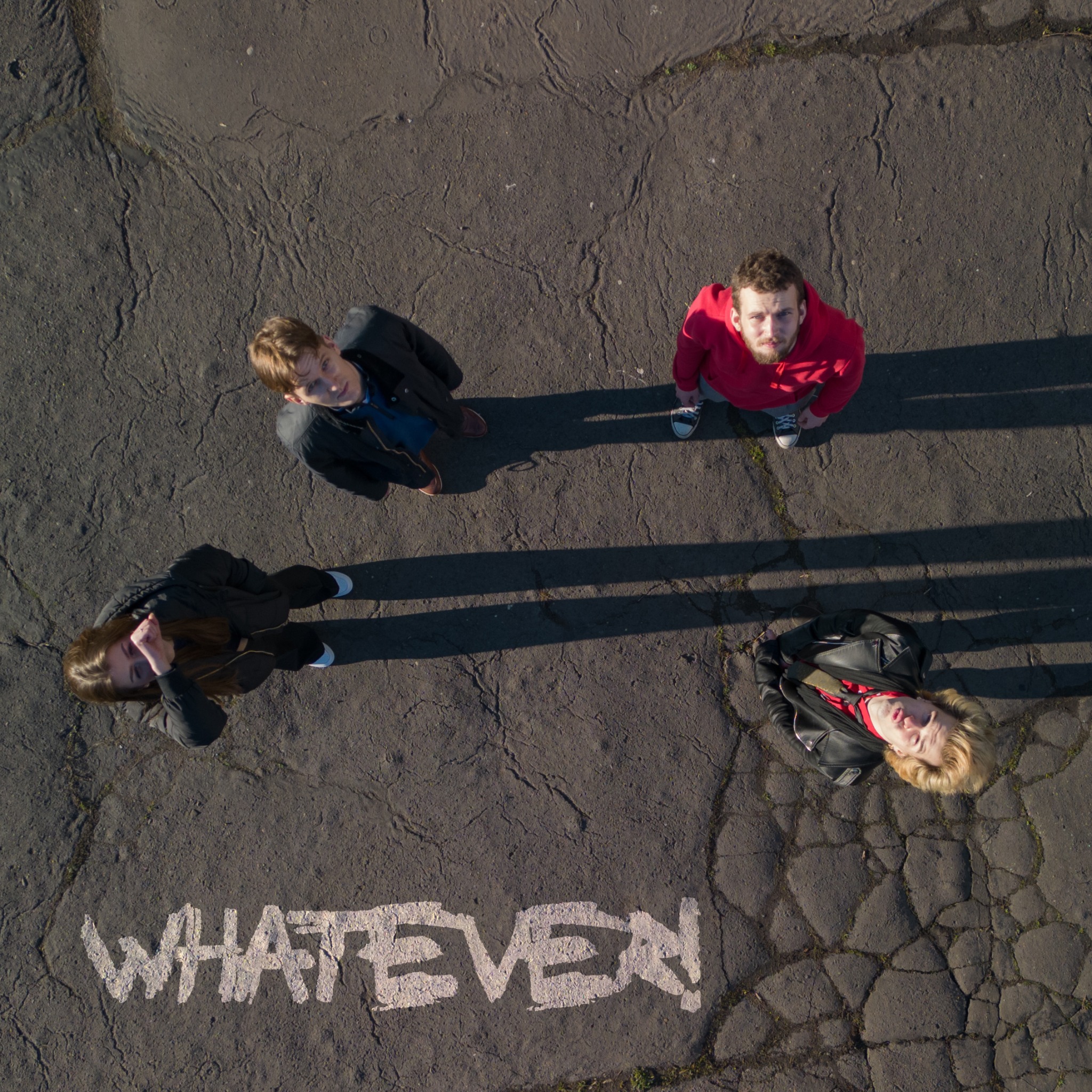 Cztery osoby stojące na popękanym asfalcie, patrzące w górę, z napisem 'WHATEVER!' namalowanym obok nich, widok z lotu ptaka.