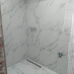 Prysznic z białymi płytkami imitującymi marmur, widoczny odpływ liniowy, bateria i fragment surowej ściany z cegły oraz siatka zabezpieczająca.