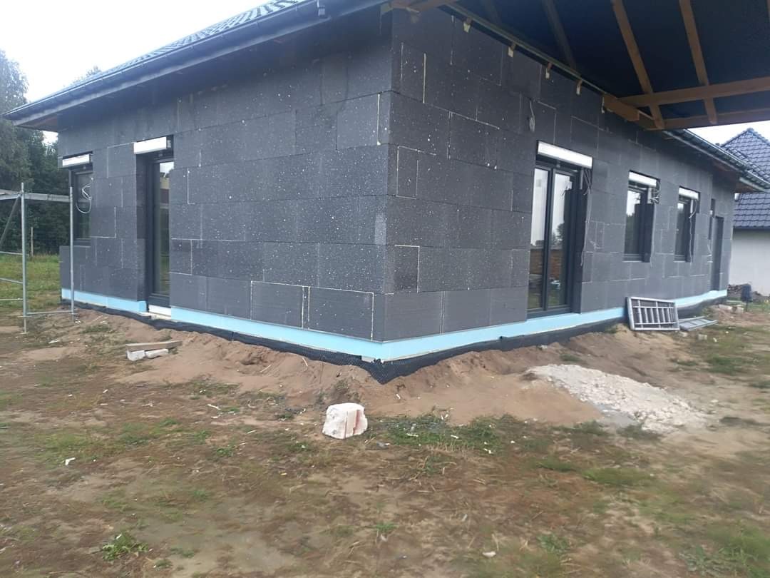 Surowy stan elewacji domu jednorodzinnego z szarego styropianu, widoczne okna z ciemnymi ramami, fragment rusztowania i niezagospodarowany teren wokół budynku.