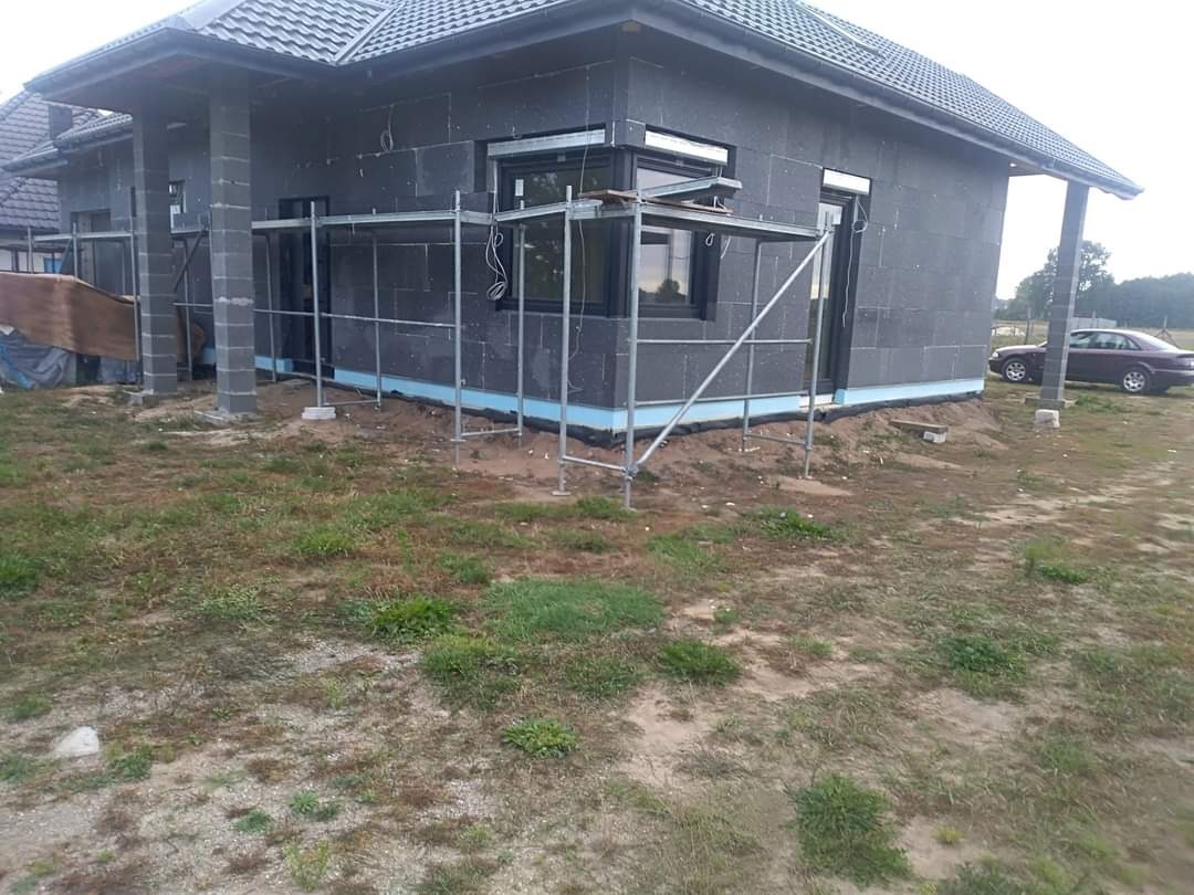 Dom w trakcie ocieplania poddasza, widoczne rusztowania, szare bloki izolacyjne, niebieska folia izolacyjna przy fundamencie i samochód osobowy w tle.