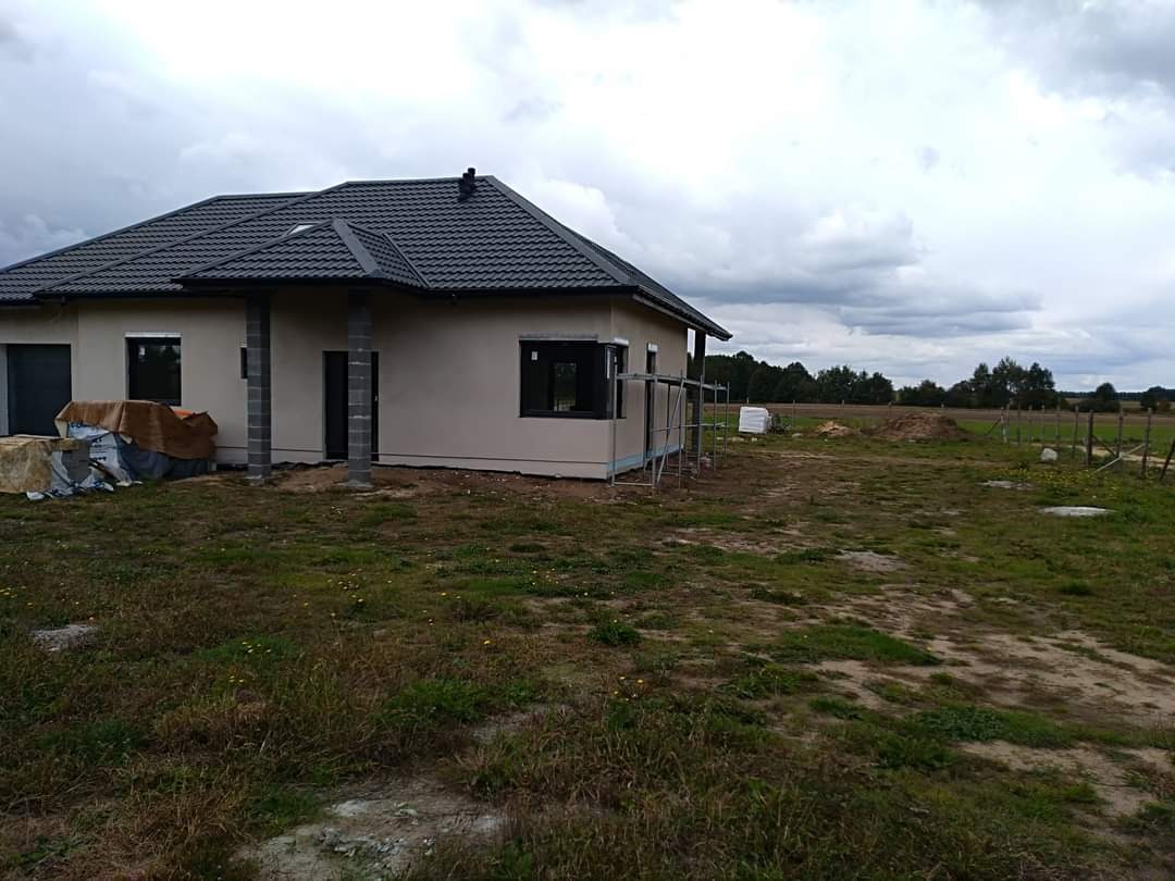 Niedawno wybudowany dom jednorodzinny z ciemną dachówką, szarymi ścianami zewnętrznymi i czarnymi ramami okiennymi, częściowo otoczony rusztowaniem, na tle pochmurnego nieba i pola.