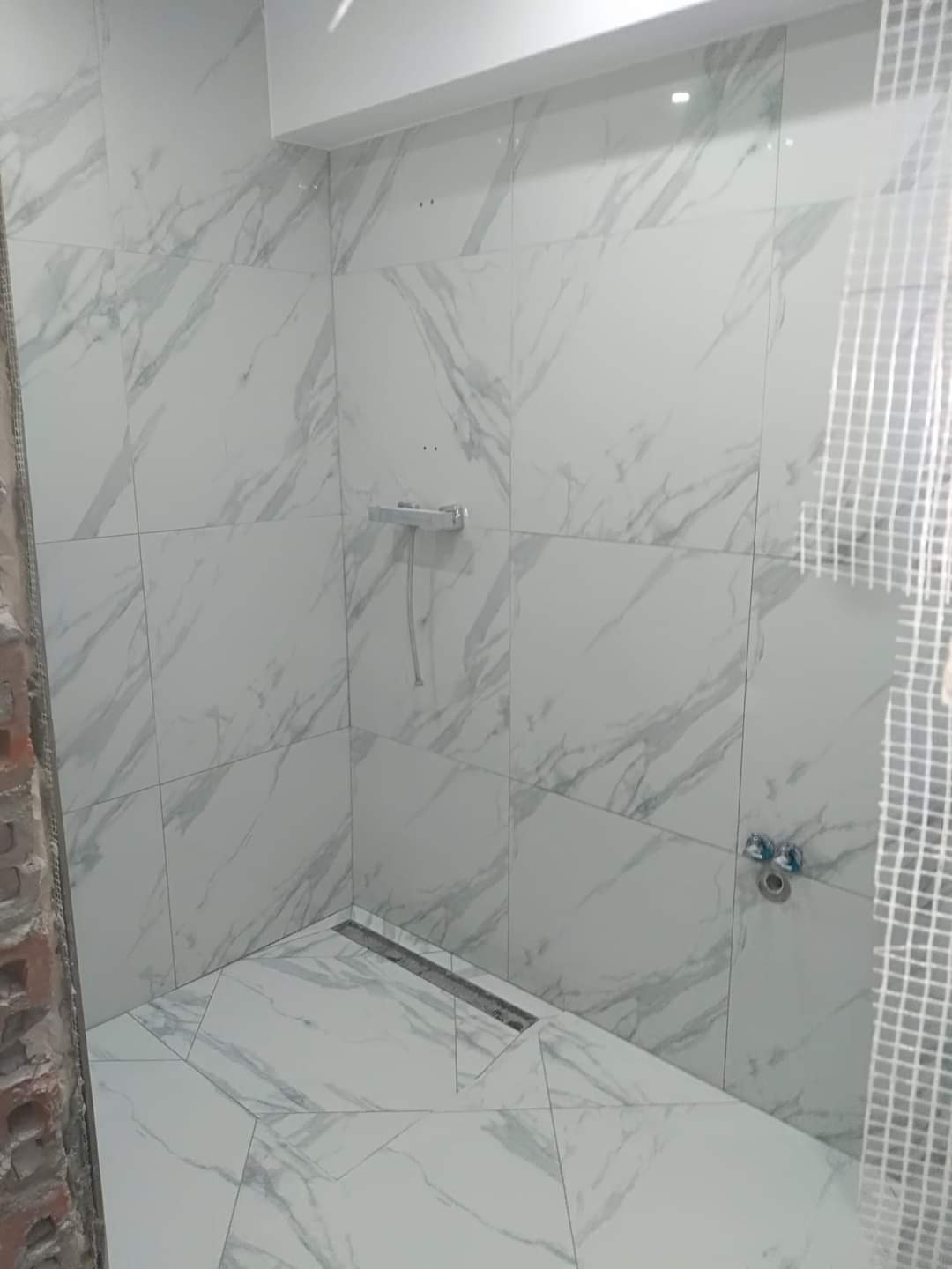 Prysznic z białymi płytkami imitującymi marmur, widoczny odpływ liniowy, bateria i fragment surowej ściany z cegły oraz siatka zabezpieczająca.