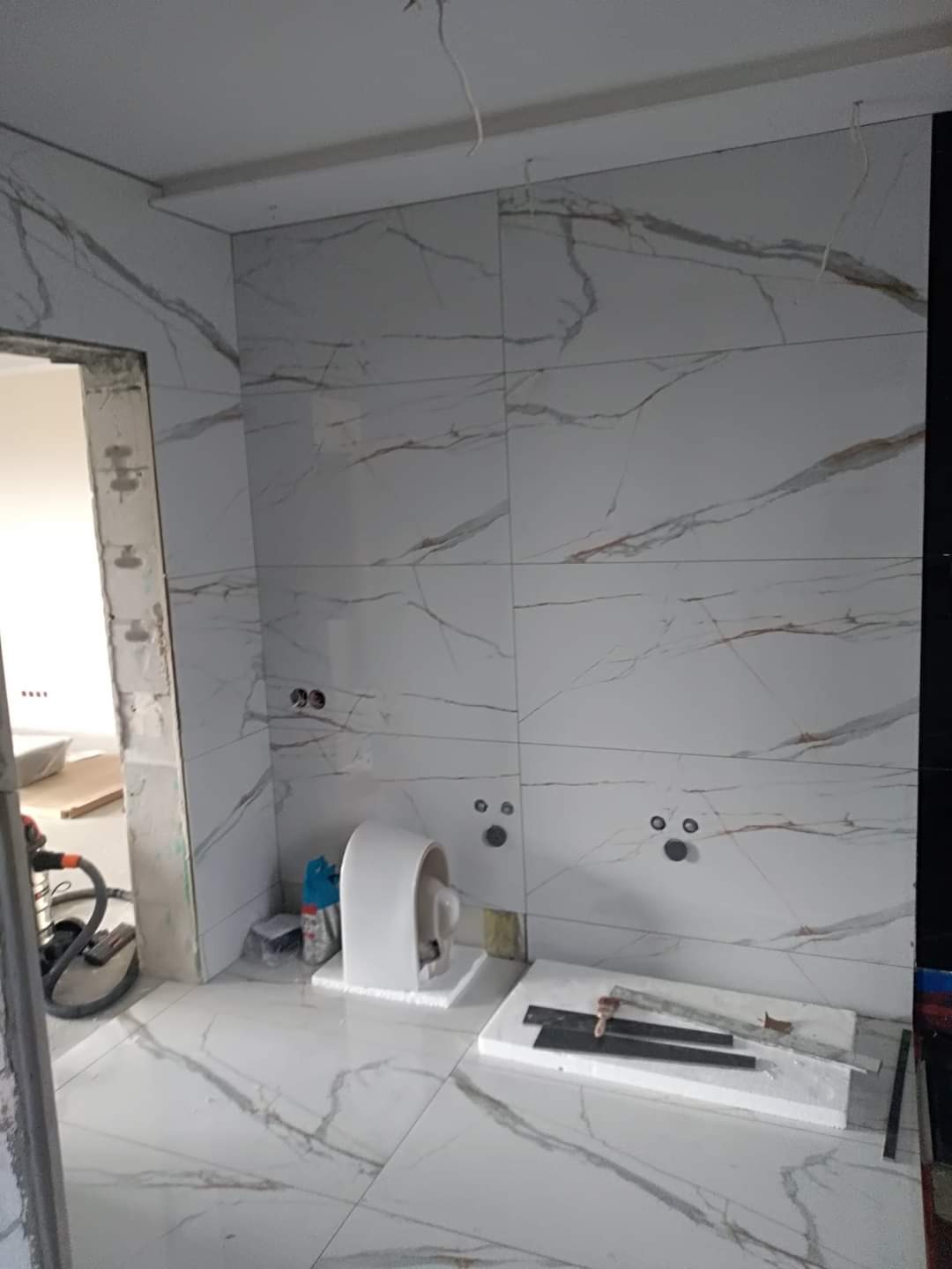 Montaż płytek imitujących marmur w łazience, widoczne otwory na instalacje, częściowo zamontowana toaleta i narzędzia glazurnicze na podłodze.