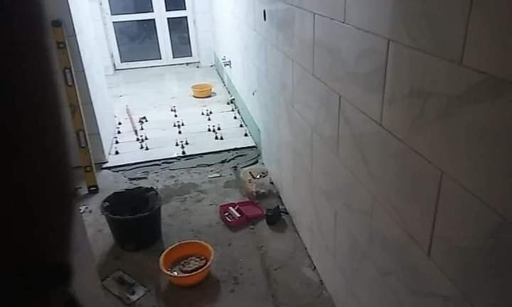 Układanie jasnych płytek na podłodze w trakcie prac, widoczne poziomica, wiadro z zaprawą, krzyżyki dystansowe i fragment ułożonej ściany z płytek.