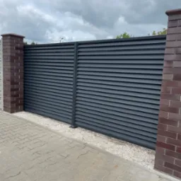 Nowoczesna, przesuwna brama wjazdowa w kolorze grafitowym z poziomymi lamelami, zamontowana między dwoma słupkami z cegły klinkierowej, widok z zewnątrz na tle pochmurnego nieba.
