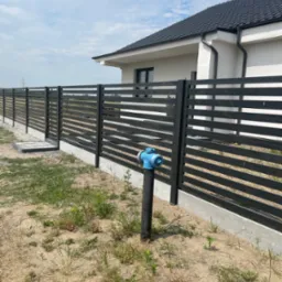 Nowoczesne, poziome, czarne ogrodzenie panelowe z furtką i betonową podmurówką w Kwilczu, widoczne na tle domu z ciemną dachówką i niebieski hydrant.