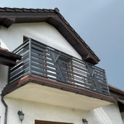 Nowoczesna, grafitowa barierka balkonowa z poziomych profili, z diagonalnymi wzmocnieniami na narożach, zamontowana na balkonie domu z białą elewacją i brązowym dachem.