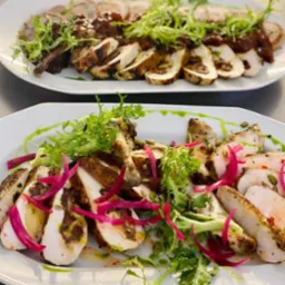 Dwa białe talerze z plastrami pieczonego mięsa, dekorowane zieleniną, czerwoną cebulą i sosem, ułożone na szarym tle.