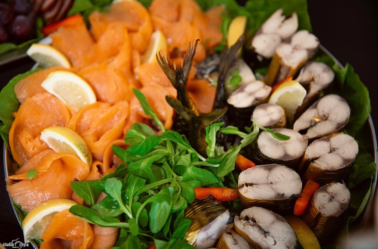 Dekoracyjnie ułożone na liściach sałaty wędzone ryby: łosoś w plastrach z cytryną oraz przekrojone makrele, ozdobione zielonymi listkami i ogonami ryb.