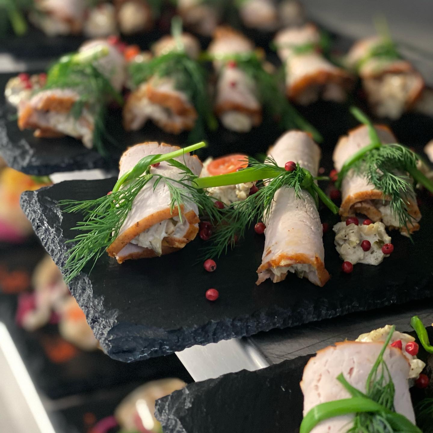 Dekoracyjnie ułożone roladki z wędliny z farszem, udekorowane koperkiem i różowym pieprzem, podane na czarnych, łupkowych talerzykach.