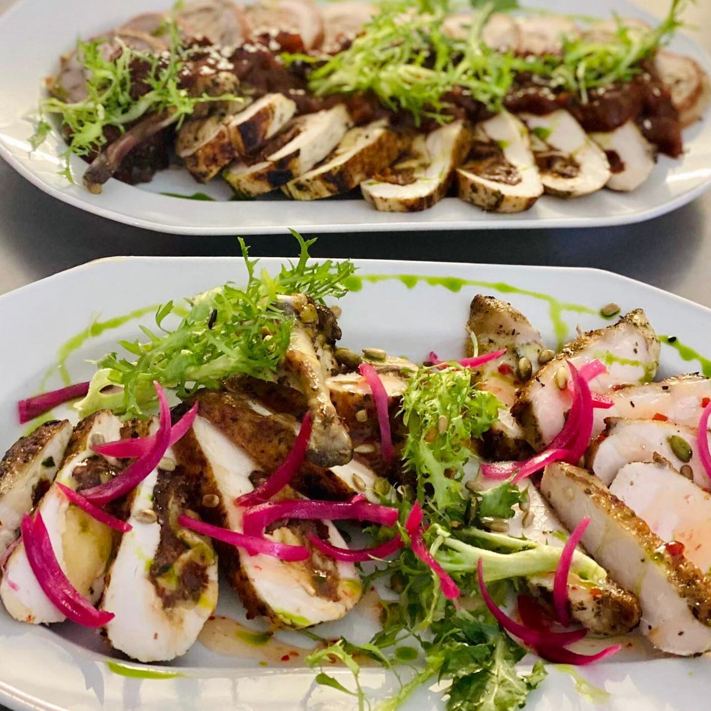 Dwa białe talerze z plastrami pieczonego mięsa, dekorowane zieleniną, czerwoną cebulą i sosem, ułożone na szarym tle.