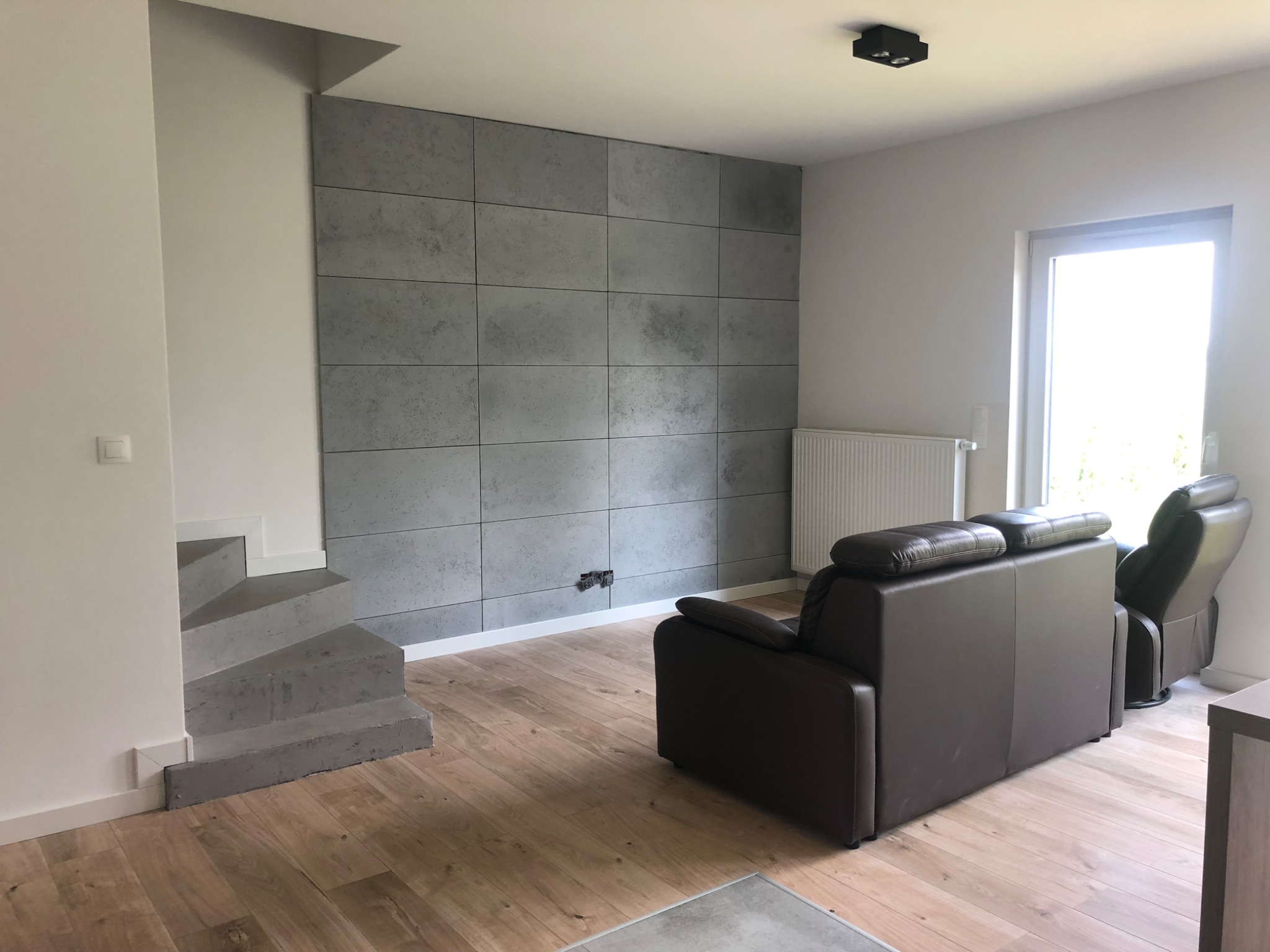 Salon z szarą ścianą wyłożoną płytami imitującymi beton, drewnianą podłogą, betonowymi schodami i brązowym skórzanym fotelem z funkcją relaks.