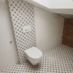 Biała toaleta wisząca w nowoczesnej łazience z ukośnym sufitem i wzorzystymi płytkami na podłodze i ścianie za toaletą.