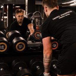 Mikołaj Biziak Trener Personalny - Mężczyzna w czarnej koszulce z napisem 'TRENER PERSONALNY MIKOLAJ BIZIAK' układa hantle na stojaku w siłowni, widoczny w odbiciu lustra