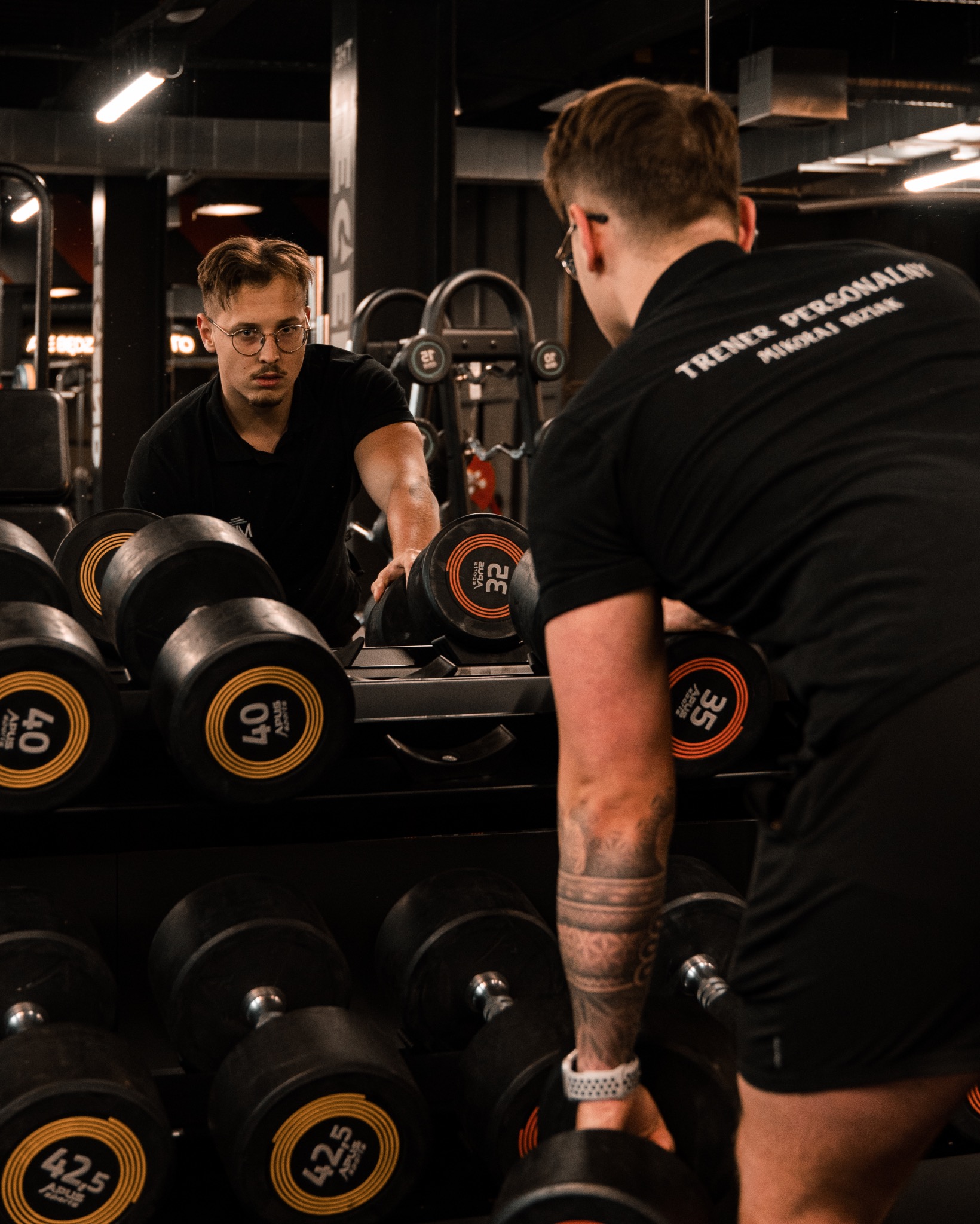Mężczyzna w czarnej koszulce z napisem 'TRENER PERSONALNY MIKOLAJ BIZIAK' układa hantle na stojaku w siłowni, widoczny w odbiciu lustra