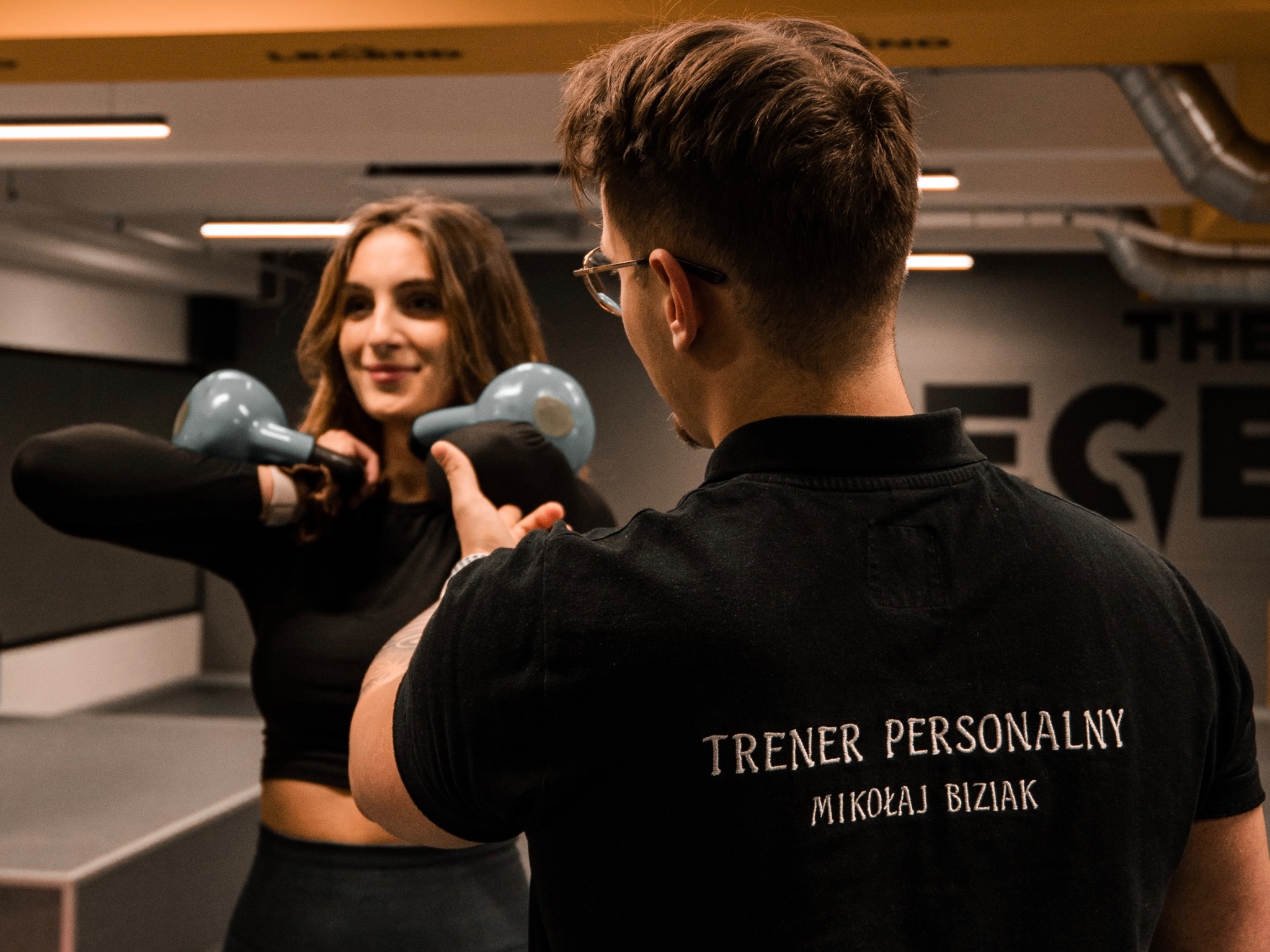 Kobieta uśmiecha się, ćwicząc z dwoma kettlebellami pod okiem mężczyzny w czarnej koszulce z napisem 'TRENER PERSONALNY MIKOŁAJ BIZIAK' na plecach, w tle siłownia.