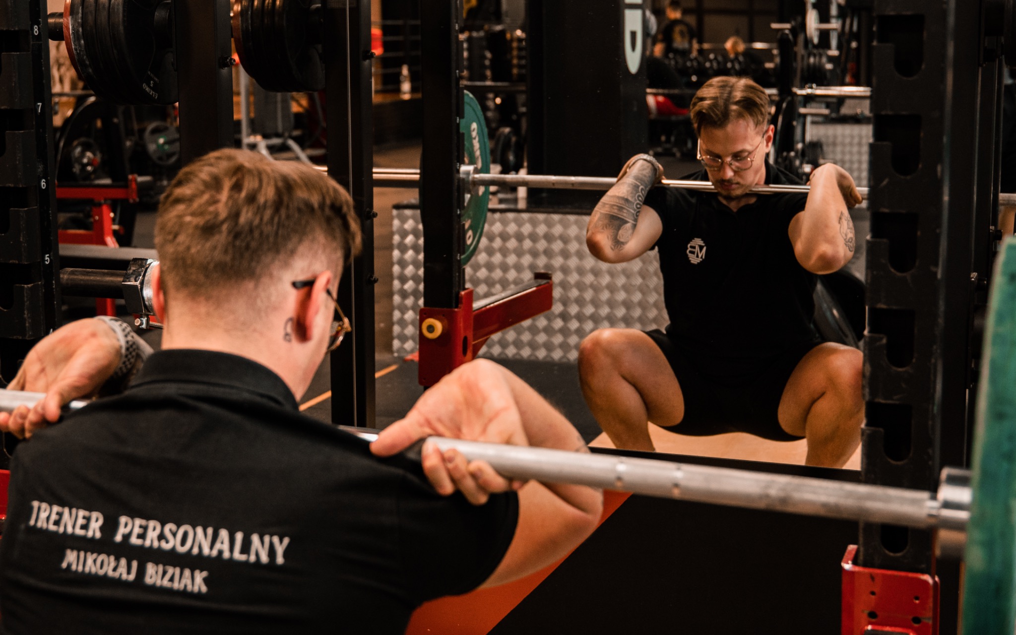 Mężczyzna w czarnej koszulce z napisem 'TRENER PERSONALNY MIKOŁAJ BIZIAK' wykonuje przysiad ze sztangą w siłowni, widoczny również jego tatuaż na ramieniu.