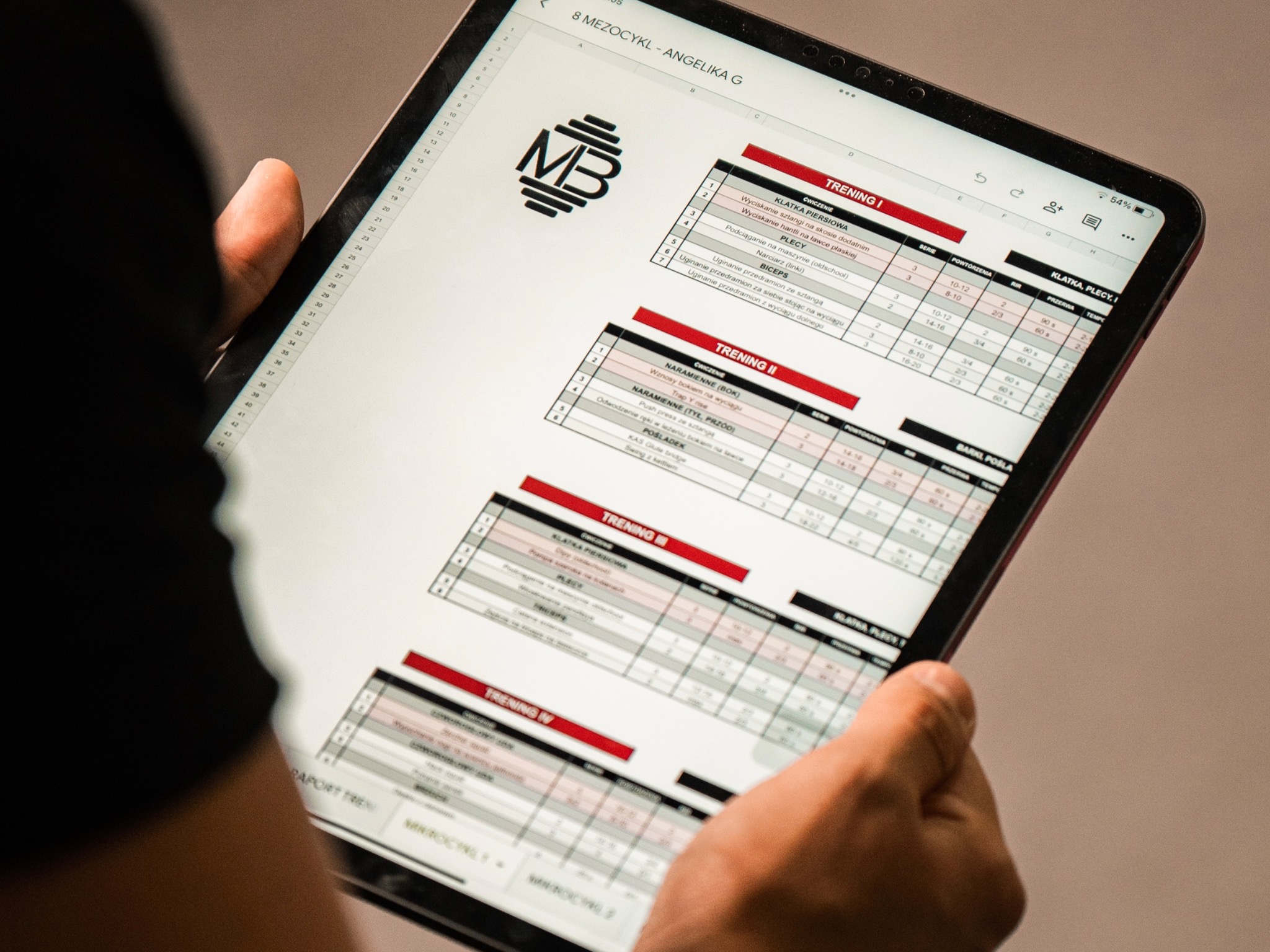 Trener personalny prezentuje plan treningowy na tablecie, szczegółowa rozpiska ćwiczeń, serii, powtórzeń i przerw, z logo MB w lewym górnym rogu.