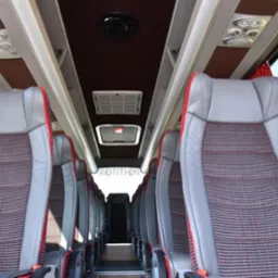 Wnętrze autobusu z rzędami szarych foteli z czerwonymi akcentami i pasami bezpieczeństwa, widok z przodu pojazdu, oświetlenie sufitowe.