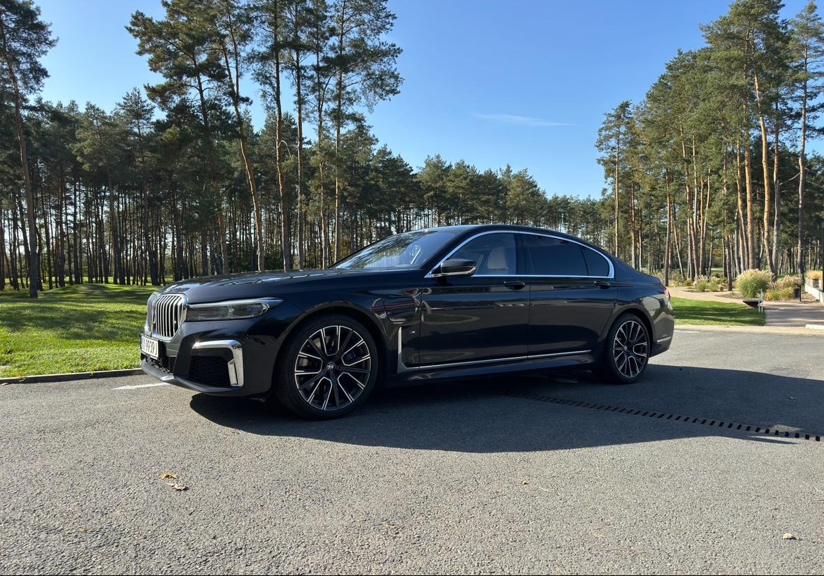 Czarna limuzyna BMW serii 7 z charakterystycznymi felgami, zaparkowana na parkingu z widokiem na las.