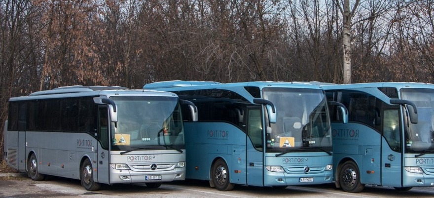 Flota autobusów firmy przewozowej, z dominującymi kolorami szarym i błękitnym, zaparkowana na tle drzew bez liści.