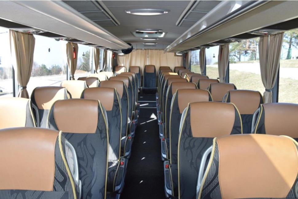 Wnętrze autobusu turystycznego z brązowymi zagłówkami, szarymi siedzeniami w czarne wzory i beżowymi zasłonami przy oknach.