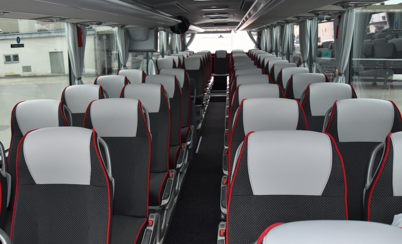 Wnętrze autobusu turystycznego z rzędami czarno-szarych siedzeń z czerwonymi akcentami, widok na przejście i okna z zasłonami.