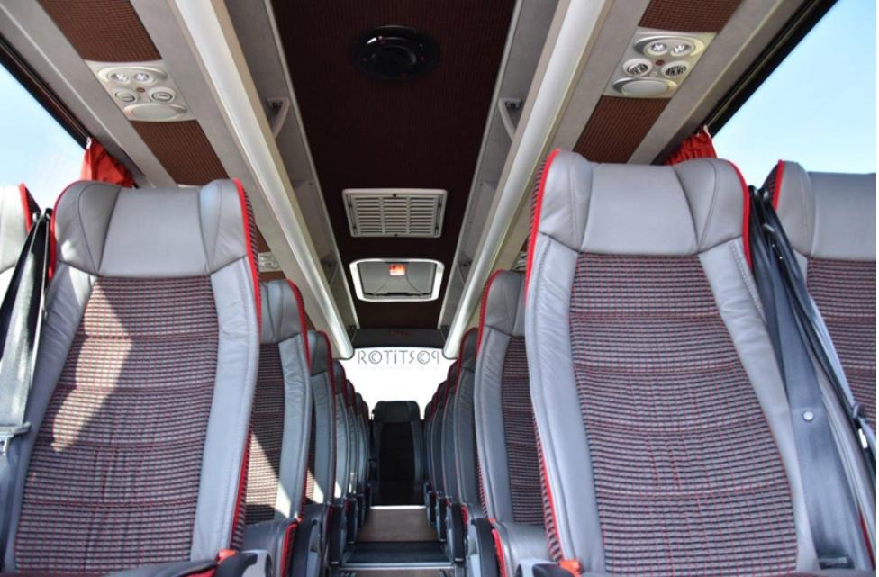 Wnętrze autobusu z rzędami szarych foteli z czerwonymi akcentami i pasami bezpieczeństwa, widok z przodu pojazdu, oświetlenie sufitowe.