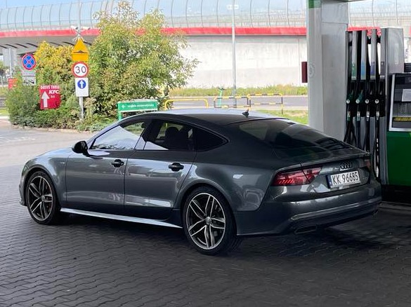 Szary samochód Audi A7 na stacji benzynowej z widoczną tablicą rejestracyjną KK 96685 i zielonymi dystrybutorami paliwa w tle.