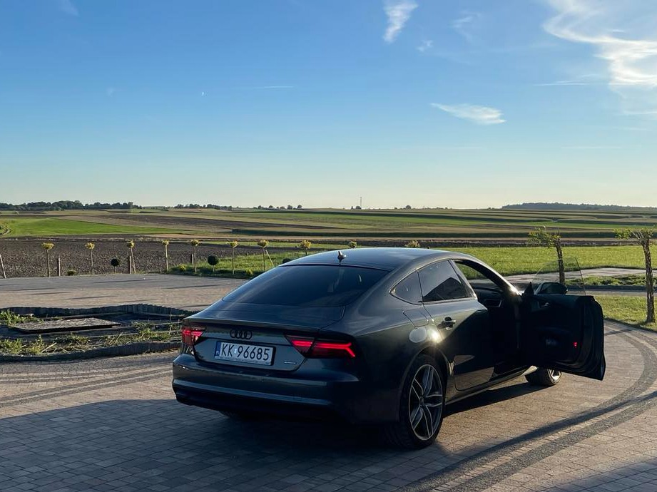 Ciemnoszary samochód Audi A7 z otwartymi drzwiami, zaparkowany na brukowanym podjeździe z widokiem na pola uprawne pod błękitnym niebem.