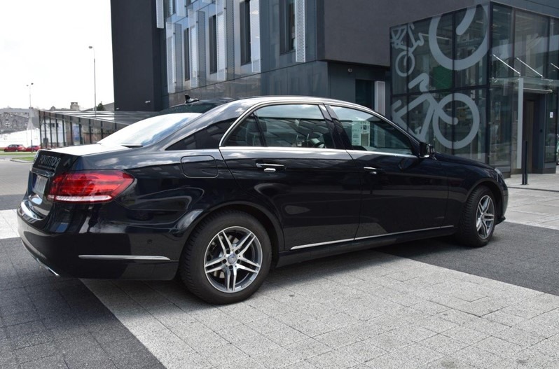 Czarny Mercedes E-klasa zaparkowany przed nowoczesnym budynkiem z szarą kostką brukową, widok z tyłu i boku.