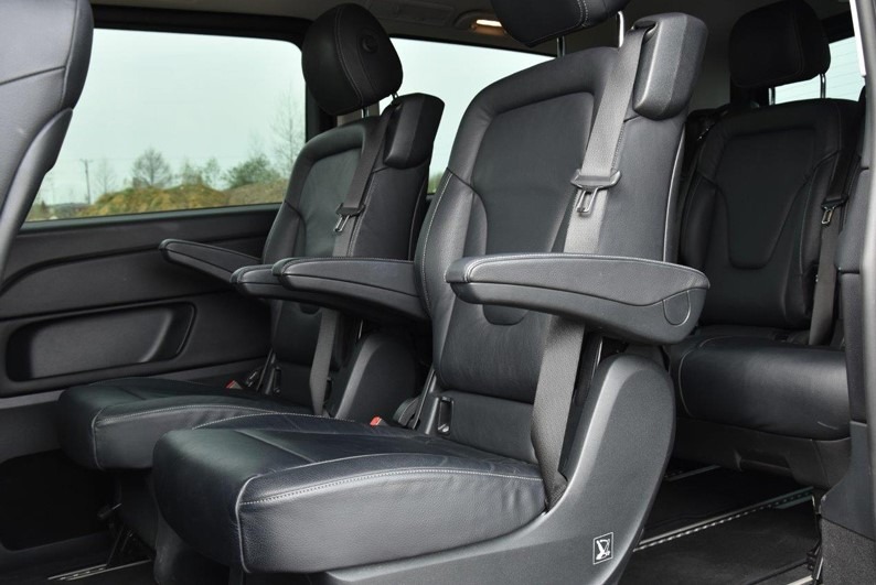 Wnętrze luksusowego busa z trzema rzędami czarnych skórzanych foteli z regulowanymi zagłówkami i podłokietnikami, widok z boku na drugi rząd foteli.