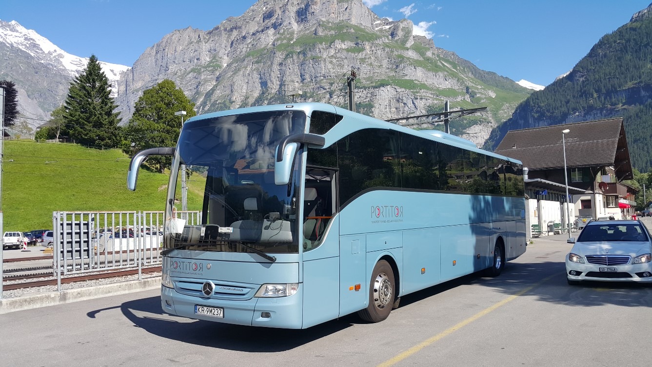Błękitny autokar turystyczny marki Mercedes z logo firmy Portitor na tle alpejskiego krajobrazu z ośnieżonymi szczytami.