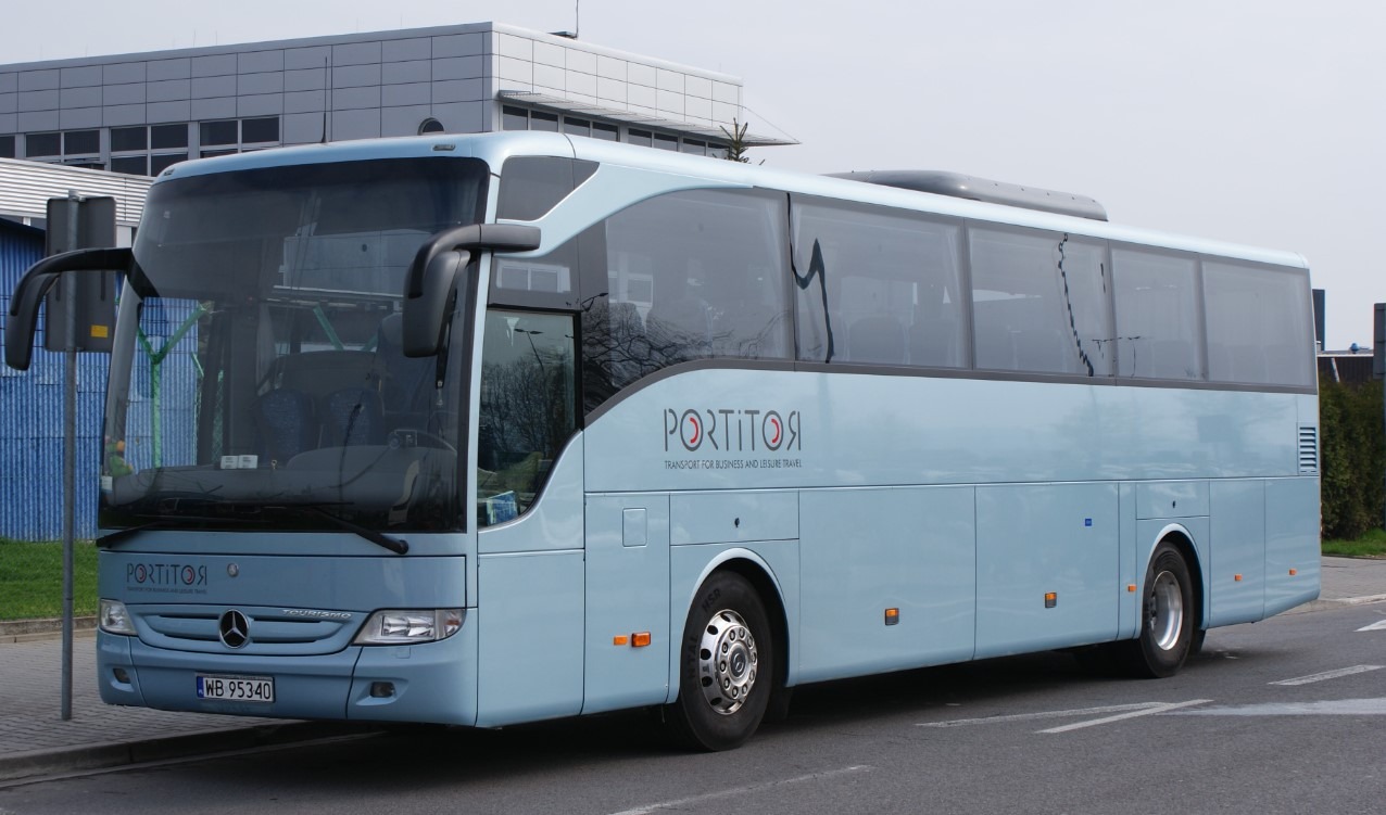 Błękitny autokar marki Mercedes z napisem 'PORTITOR' na tle budynku biurowego i zieleni.