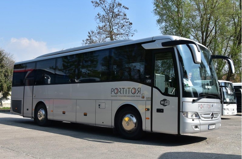 Nowoczesny autokar marki Mercedes w kolorze srebrnym z napisem 'PORTITOR' na boku, zaparkowany na placu w słoneczny dzień.